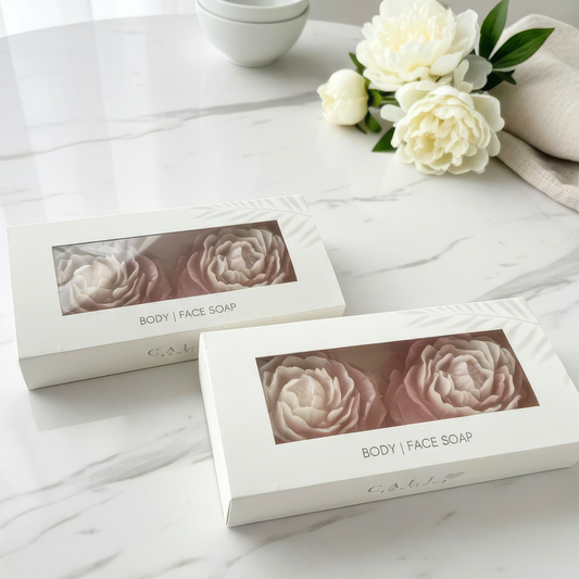 Two linen branded boxes of body and face soap with peony-shaped soaps on a marble surface.