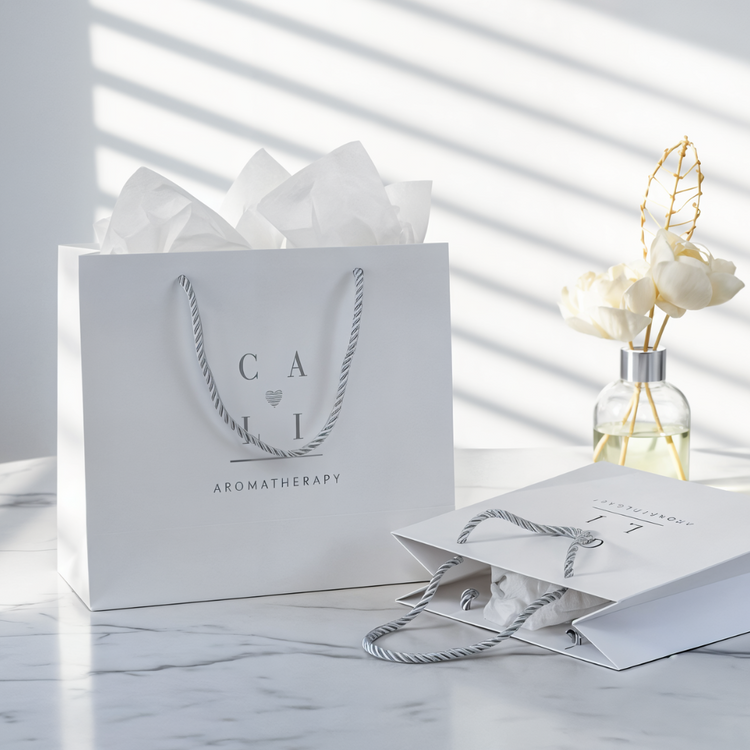 two boutique white shopping bags with light gray branding and silver twisted handles and reed diffuser in the background on a marble surface.