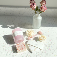 iridescent Pink candle, soap, and heart shaped coaster items on a textured surface with a vase of pink roses in the background.