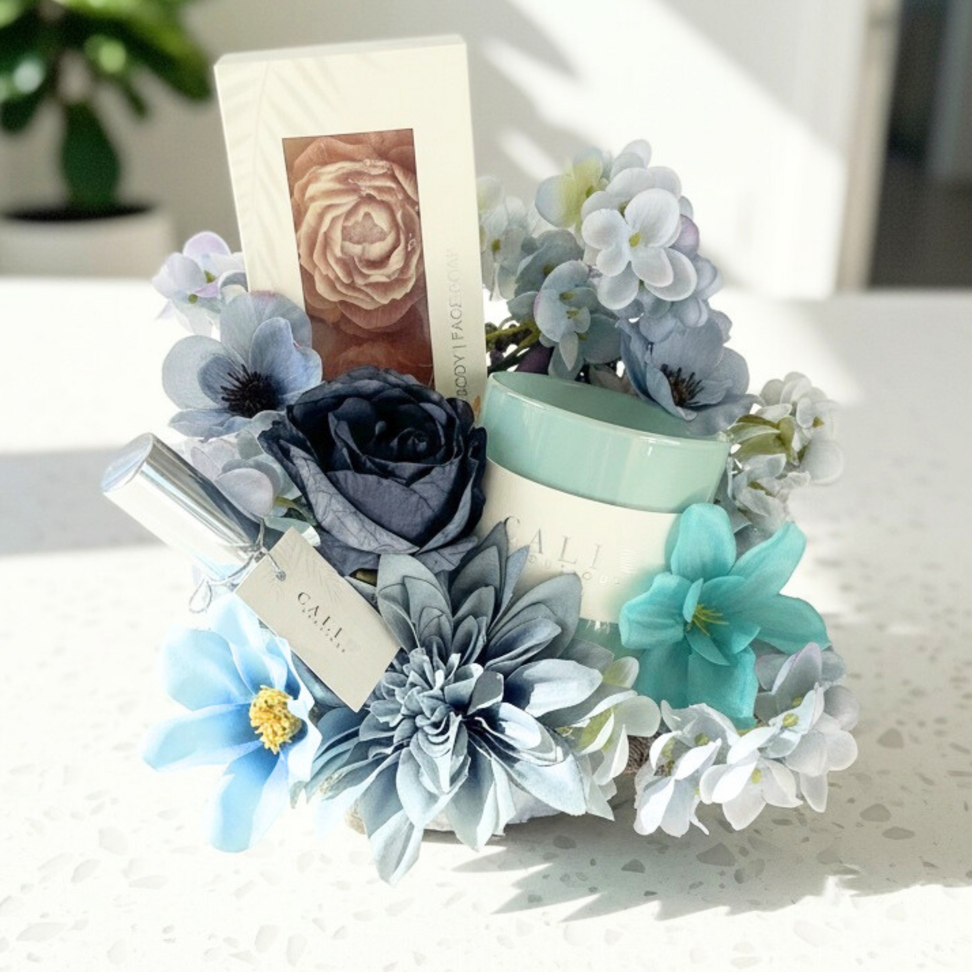 Decorative floral arrangement of blue flowers with a candle, soap set and mood mist spray on a white surface.