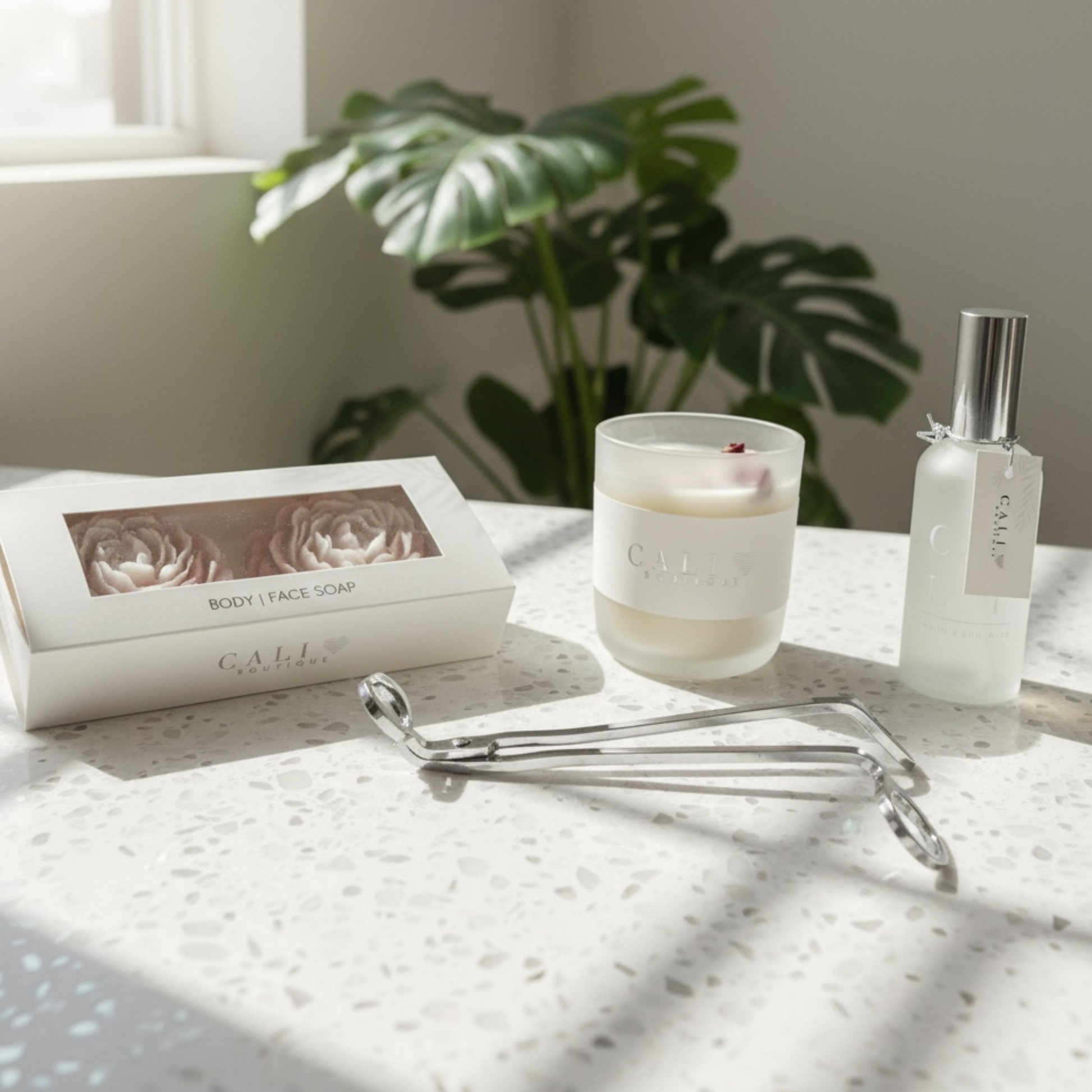 Candle, soap, body mist, and wick trimmer on a marble surface with a plant in the background