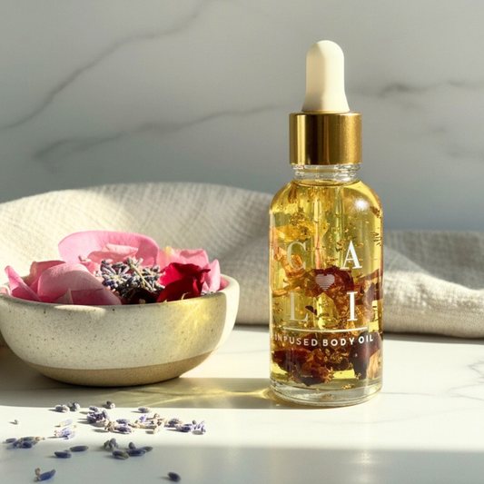 Bottle of infused body oil with a gold dropper with rose petals and 24k gold next to a bowl of flowers on a marble surface.