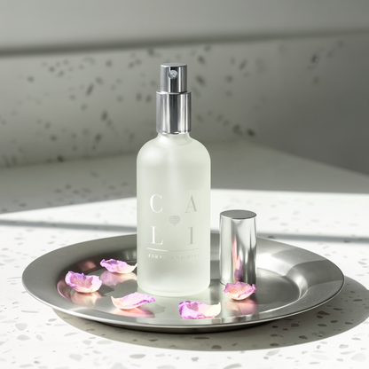 Frosted spray bottle labeled 'CALI Love Aromatherapy on a silver tray with pink petals, against a light background.